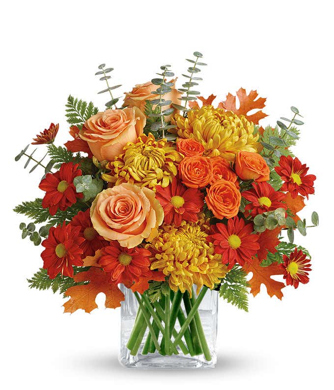 A stunning floral arrangement featuring deluxe and premium orange roses, orange spray roses, bronze mums, and red daisies, accented with spiral eucalyptus, all elegantly displayed in a clear cube vase.