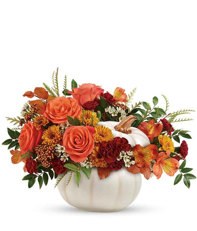 A charming floral arrangement featuring orange roses, orange alstroemeria, maroon mini carnations, yellow mums, and white waxflower, all beautifully arranged in a ceramic pumpkin.