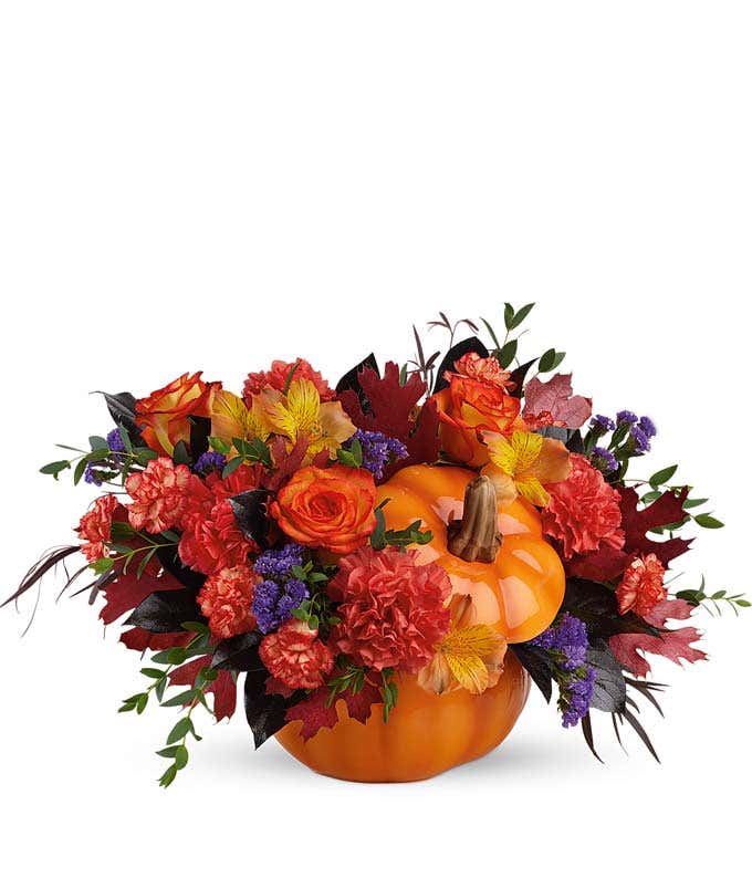 A vibrant floral arrangement featuring orange carnations, orange mini carnations, orange Peruvian lilies, purple statice, deluxe and premium bi-color roses, and oak leaves, all beautifully presented in a keepsake container.