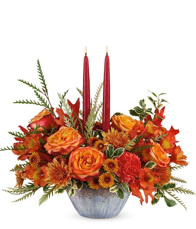  A vibrant autumn-themed floral arrangement featuring orange fall roses, orange alstroemeria, orange carnations, orange button mums, bronze cushion mums, and oak leaves, complemented by taper candles, all beautifully presented in a keepsake bowl.