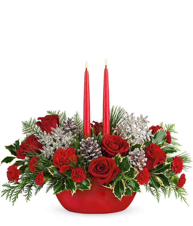 A festive arrangement with red roses, red carnations, red mini carnations, silver snowflakes, snowy pinecones, evergreens, and red candles, displayed in a keepsake container.