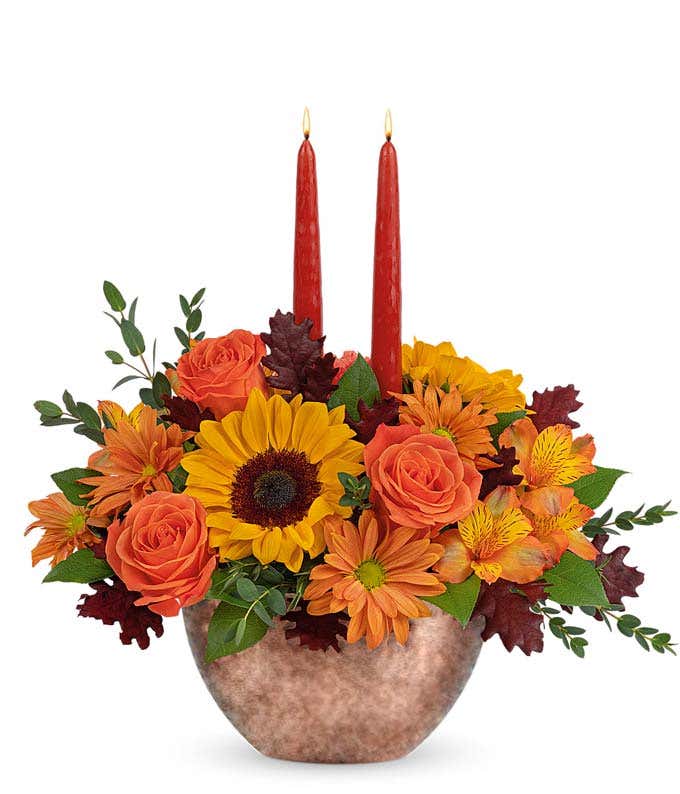  A cheerful floral arrangement featuring sunflowers, orange carnations, bronze daisies, orange alstroemeria, and seasonal greenery, complemented by taper candles, all beautifully presented in a keepsake bowl.