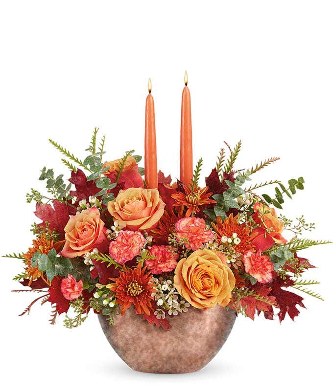 A warm and inviting floral arrangement featuring orange roses, orange mini carnations, bronze mums, and white waxflower, accented with oak leaves and seasonal greenery, complemented by taper candles, all beautifully presented in a keepsake bowl.