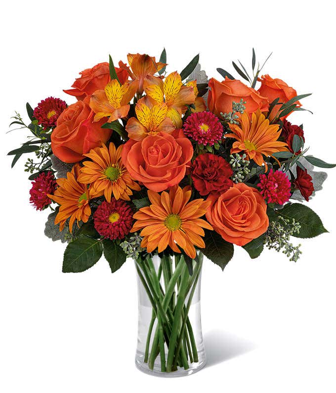 A vibrant floral arrangement featuring orange roses, orange alstroemeria, bronze daisies, red Matsumoto asters, maroon mini carnations, and seasonal greenery, complemented by a personal card message, all beautifully presented in a clear cylinder vase.