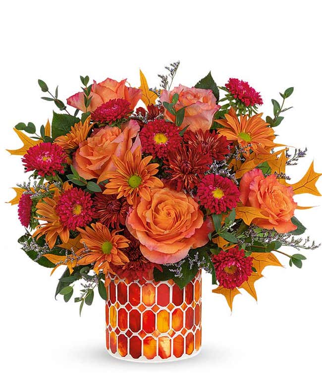 A vibrant floral arrangement featuring orange roses, red Matsumoto asters, bronze mums, and bronze daisies, complemented by greenery and fall leaves. The arrangement includes a personal card message and is elegantly presented in a keepsake vase&amp;#8203;