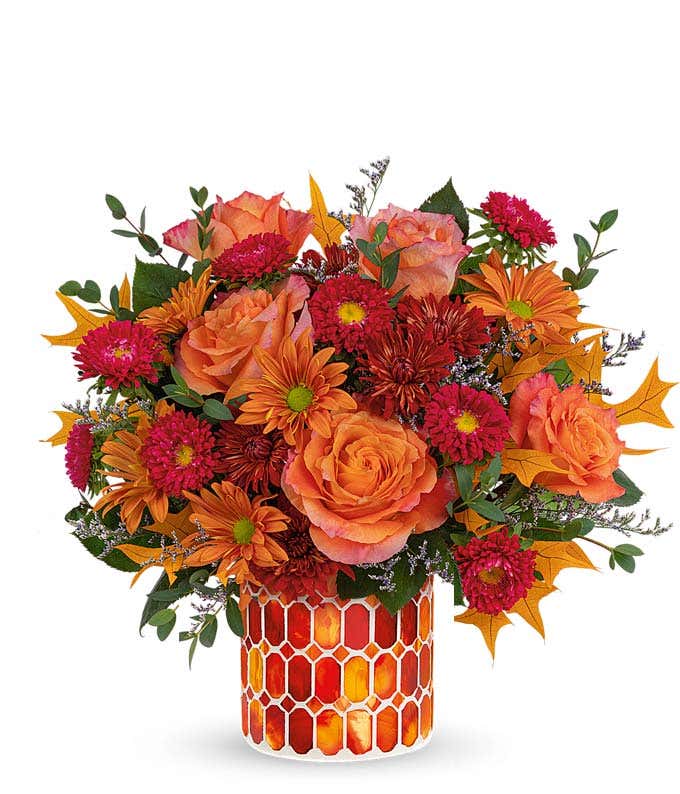 A vibrant floral arrangement featuring orange roses, red Matsumoto asters, bronze mums, and bronze daisies, complemented by greenery and fall leaves. The arrangement includes a personal card message and is elegantly presented in a keepsake vase​