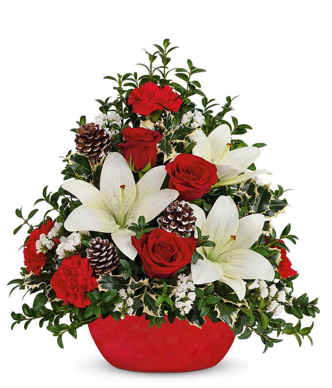 A festive arrangement with red carnations, red roses (deluxe & premium only), white lilies, white sinuata, seasonal greens, a mini Christmas tree, displayed in a red stoneware container