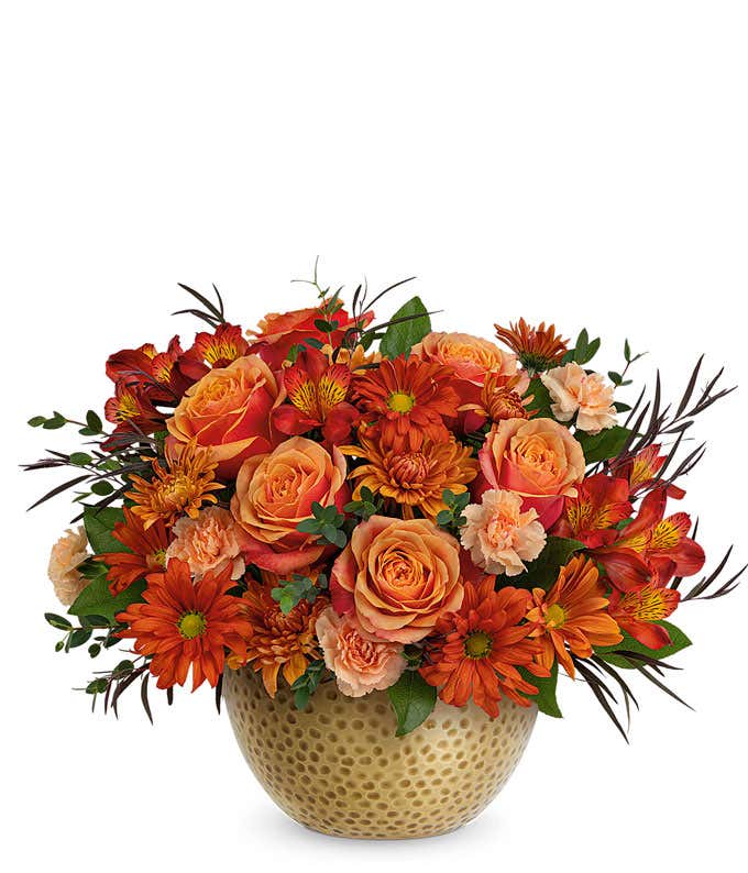 A vibrant floral arrangement featuring orange roses, red alstroemeria, peach miniature carnations, bronze cushion spray chrysanthemums, bronze daisy spray chrysanthemums, lemon leaf, parvifolia eucalyptus, and red agonis, all elegantly arranged in a brass