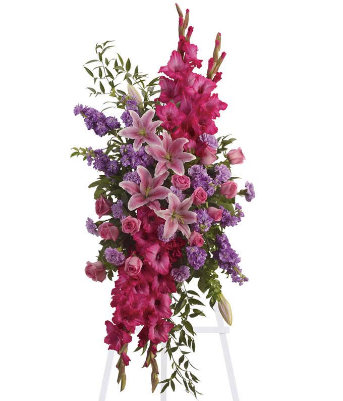 Standing spray with pink gladiolus, lilies, roses, carnations, and green foliage on a white metal easel.