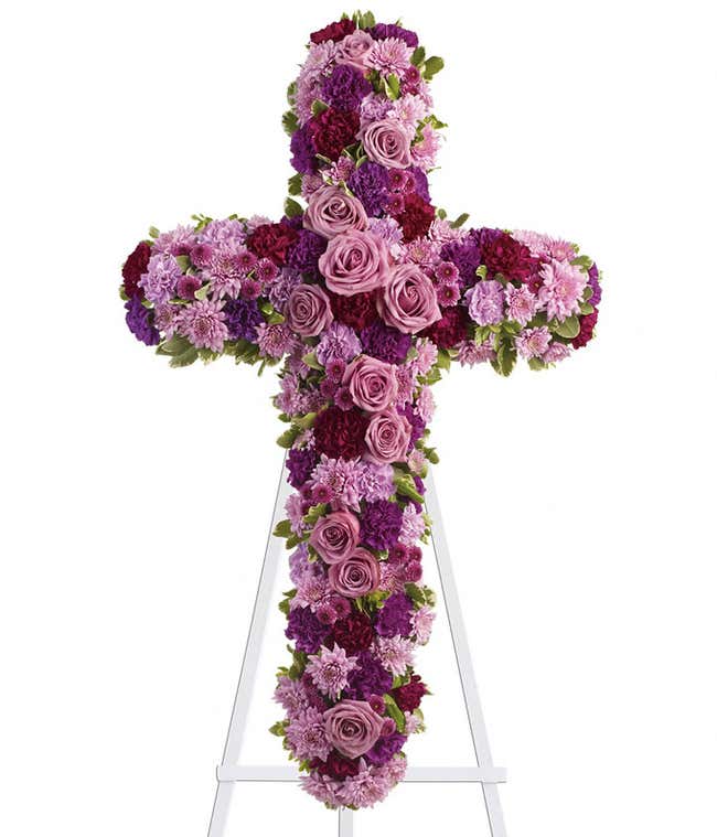 Cross-shaped standing spray with pink and purple roses, chrysanthemums, carnations, and green foliage on a white metal easel.