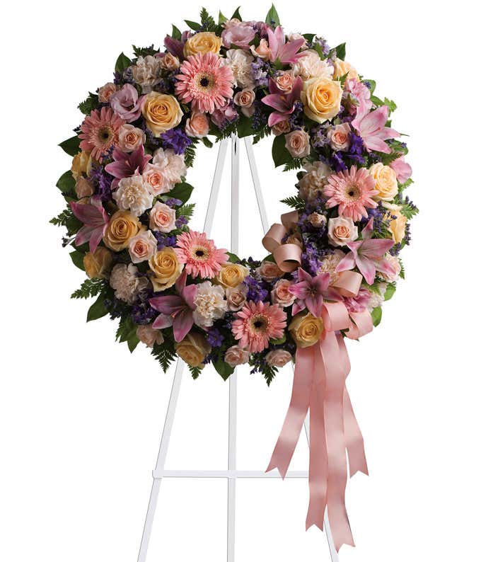 Funeral wreath with peach roses, pink lilies, peach carnations, green foliage, and a pink satin ribbon on a white metal easel.