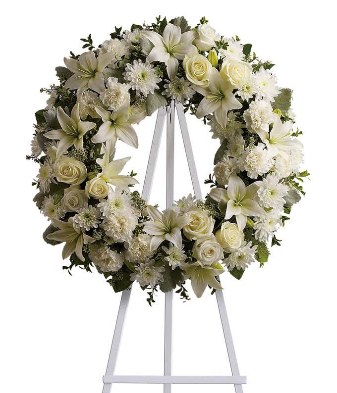 Funeral wreath with white roses, lilies, chrysanthemums, and green foliage on a white metal easel.