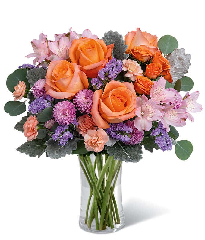 Short, round arrangement of orange roses and purple flowers, in a Tall Glass Cylinder Vase