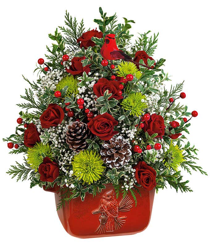 A festive arrangement with miniature red carnations, red roses, green cushion spray chrysanthemums, floral greens, displayed in a keepsake red ceramic cardinal bowl.