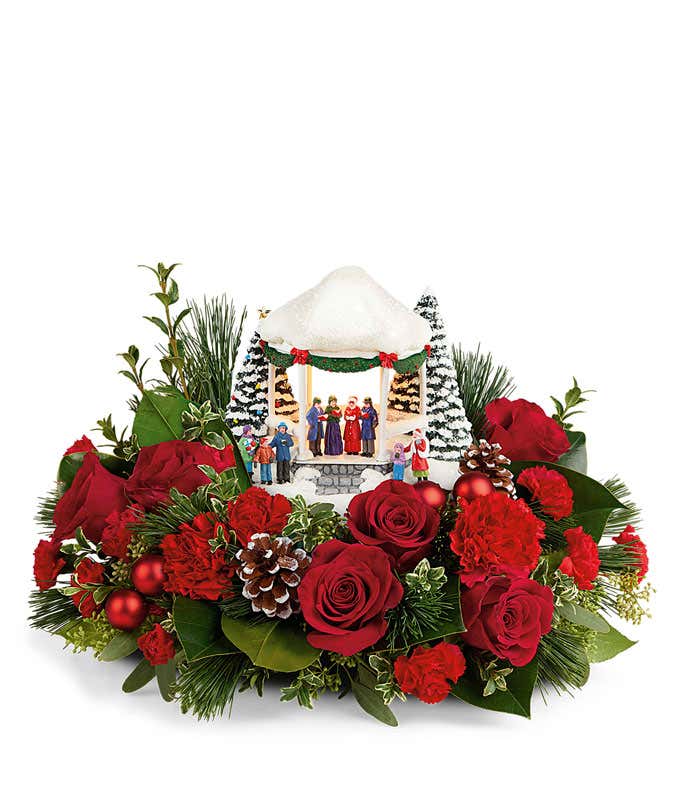 A festive arrangement with red roses, red carnations, floral greens, pinecones, and red ornaments, featuring a keepsake Thomas Kinkade Carolers on the Green sculpture.