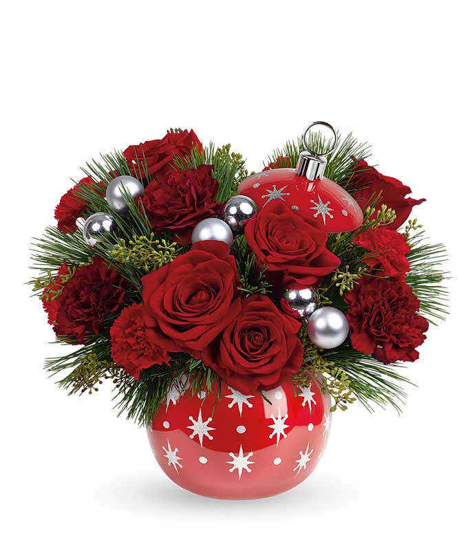 A festive arrangement with red roses, red mini carnations, seeded eucalyptus, floral greens, displayed in a keepsake red ceramic ornament vase.