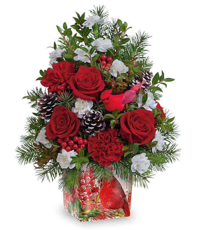 A festive arrangement with deluxe and premium red roses, premium red spray roses, red carnations, white miniature carnations, seasonal floral greens, cardinal and pinecone details, displayed in a keepsake cardinal cylinder vase.