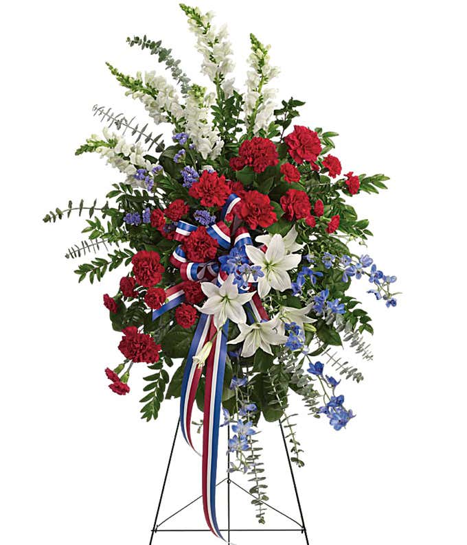 White lilies, white snapdragons, blue delphinium, red carnations with multicolored patriotic ribbon.