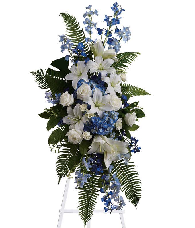 Standing spray with white lilies, roses, blue delphiniums, and green ferns on a white metal easel.