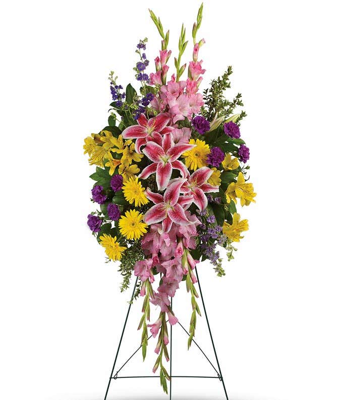 A standing funeral spray adorned with Pink Stargazer Lilies, Yellow Gerberas, Yellow Alstroemeria, Pink Gladioli, Purple Carnations, and Purple Larkspur, evoking a sense of grace, warmth, and reverence in remembrance of a beloved soul.