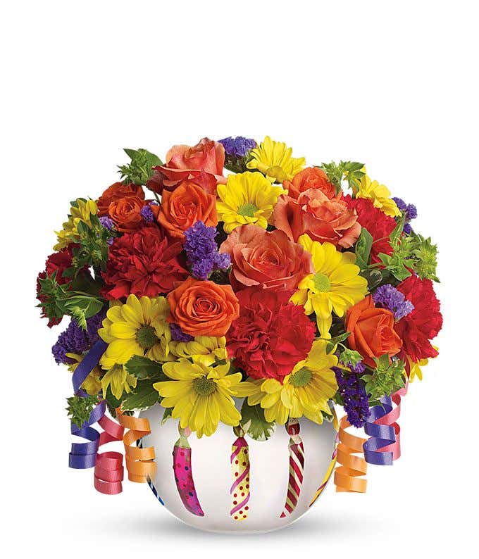 A festive arrangement featuring orange spray roses, red mini carnations, and yellow daisy spray mums, presented in a birthday cake inspired vase. This lively mix offers a fun and celebratory display.