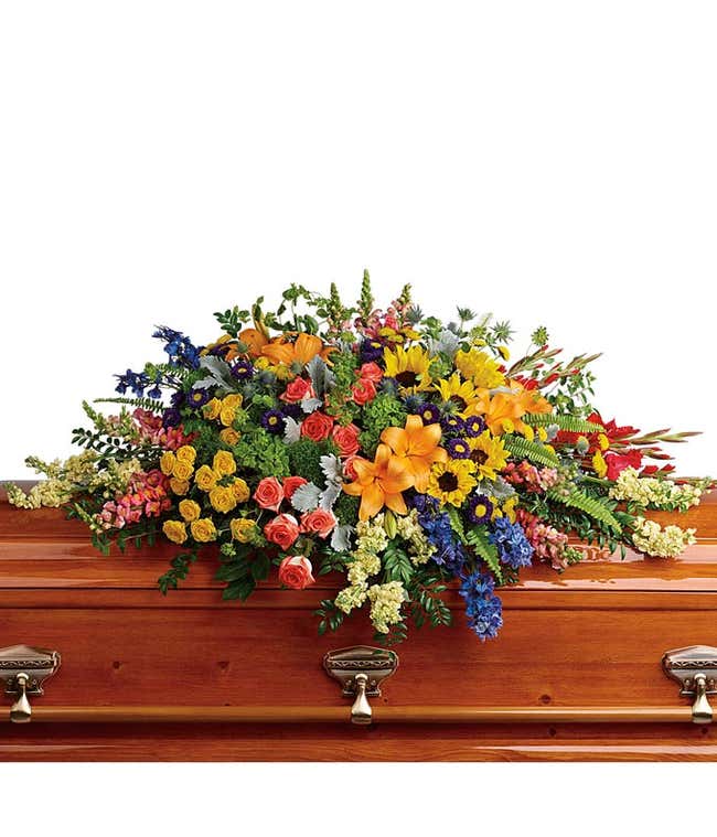 A vibrant casket spray featuring Green Hydrangea, Orange Roses, Yellow Spray Roses, Orange Asiatic Lilies, Red Gladioli, Yellow Sunflowers, Blue Delphinium, Orange Snapdragons, Light Yellow Stock Flowers, Purple Matsumoto Asters, Yellow Button Chrysanthem