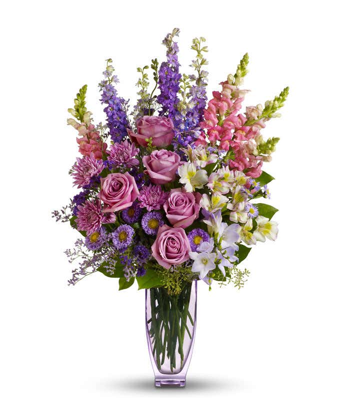 A beautiful lavender and pink arrangement in a lavender vase featuring roses, alstroemeria, snapdragons, larkspur, freesia, matsumoto asters, monte cassino asters, cushion spray mums, statice, and limonium. This elegant display offers a harmonious and soo