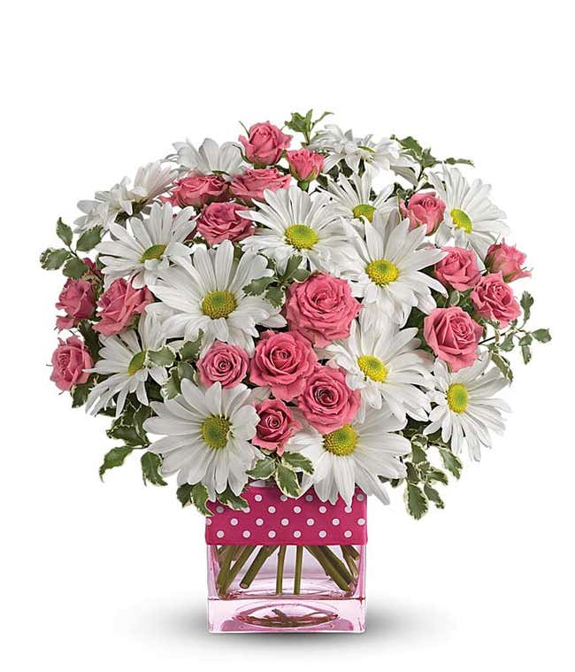 A floral arrangement featuring hot pink roses and white daisies in a pink cube vase, adorned with a polka dot ribbon.