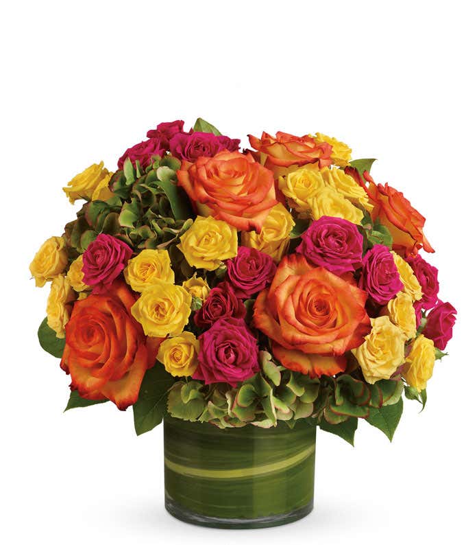 A floral arrangement featuring green hydrangea, hot pink roses, yellow roses, and bi-color roses in a clear glass vase.