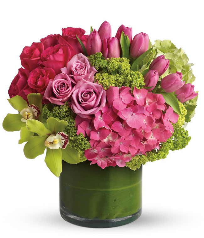 A floral arrangement featuring green cymbidium orchids, purple roses, hot pink roses, pink tulips, green hydrangea, pink hydrangea, and viburnum in a glass cylinder vase.