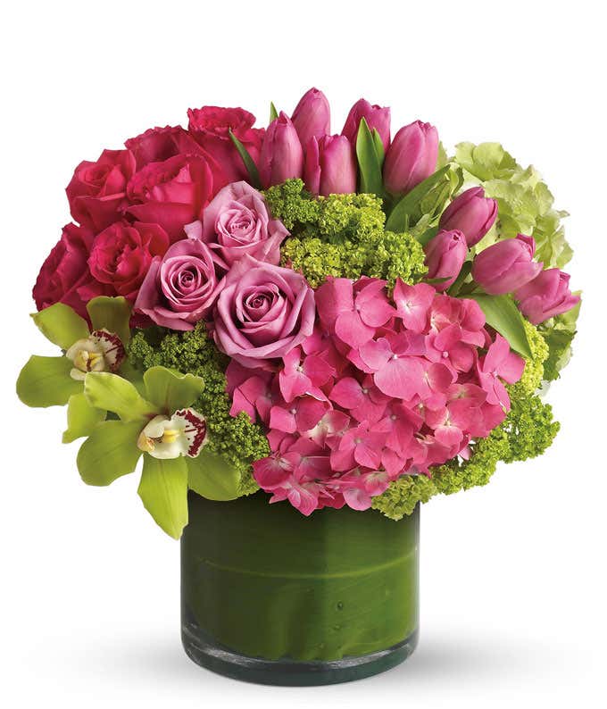 A floral arrangement featuring green cymbidium orchids, purple roses, hot pink roses, pink tulips, green hydrangea, pink hydrangea, and viburnum in a glass cylinder vase.