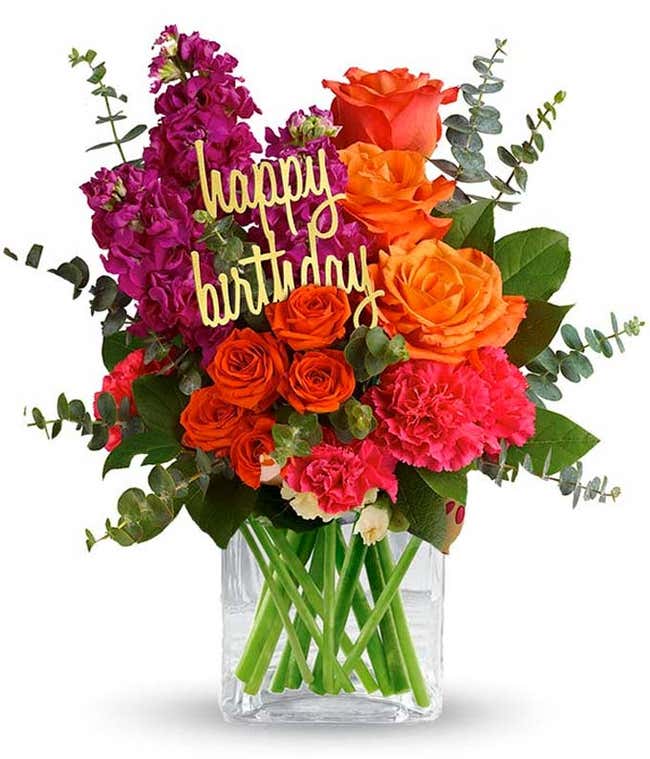 Floral arrangement featuring orange roses, orange spray roses, fuchsia stock, pink carnations, spiral eucalyptus, and lemon leaf with a 'Happy Birthday' pick in a clear cube vase.