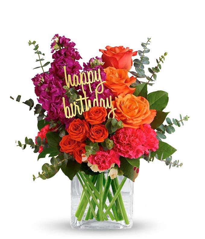 Floral arrangement featuring orange roses, orange spray roses, fuchsia stock, pink carnations, spiral eucalyptus, and lemon leaf with a 'Happy Birthday' pick in a clear cube vase.