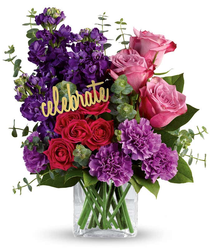 A floral arrangement featuring lavender roses, hot pink spray roses, purple stock, lavender carnations, spiral eucalyptus, and lemon leaf in a glass cube, with a 'Celebrate' pick.