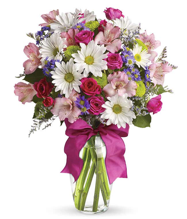 A cheerful floral arrangement featuring pink spray roses, white daisies, green button poms, purple Monte Casino, and limonium, all beautifully arranged in a glass vase with a hot pink ribbon. This vibrant mix offers a playful and lively display.