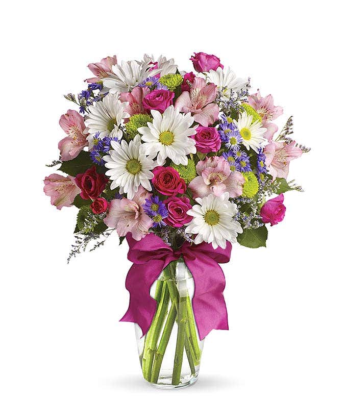 A cheerful floral arrangement featuring pink spray roses, white daisies, green button poms, purple Monte Casino, and limonium, all beautifully arranged in a glass vase with a hot pink ribbon. This vibrant mix offers a playful and lively display.