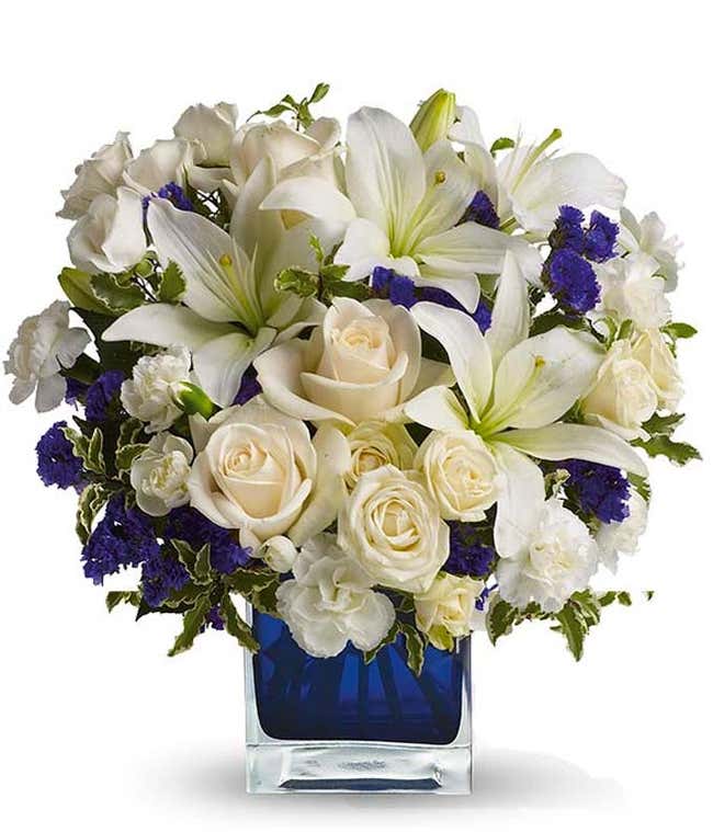 Floral arrangement featuring cr&egrave;me roses, cr&egrave;me spray roses, white Asiatic lilies, white miniature carnations, and purple statice in a blue cube vase.