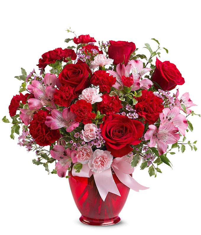 A striking arrangement featuring red roses, pink alstroemeria, red carnations, and pink waxflower, elegantly presented in a red vase with a ribbon. This bold mix offers a stunning and romantic display.