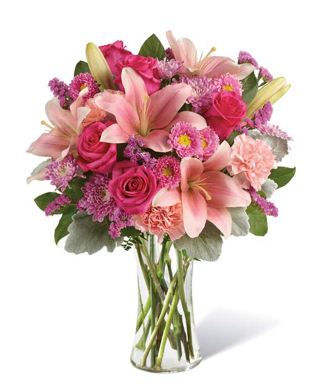 Floral arrangement featuring pink roses, pink Asiatic lilies, pink carnations, pink Matsumoto asters, lavender cushion spray chrysanthemums, pink statice, dusty miller, lemon leaf, in a clear vase.