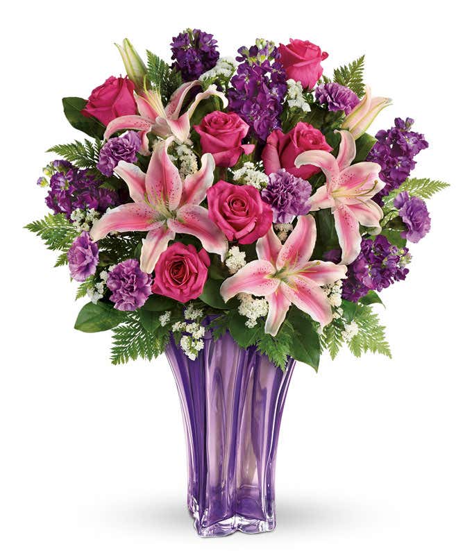 A floral arrangement featuring hot pink roses, pink oriental lilies, purple carnations, and floral greens in a keepsake lavender vase.