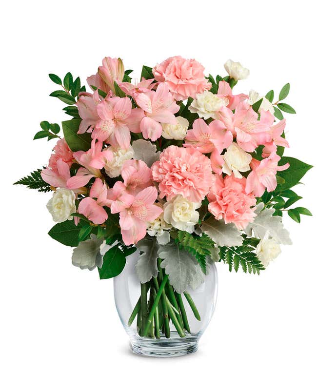 A delicate floral arrangement of pink alstroemeria, pink carnations, white mini carnations, dusty miller, huckleberry, leatherleaf fern, and lemon leaf, elegantly arranged in a clear glass vase. This soft blend offers a gentle and inviting display.