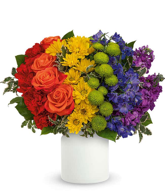 A vibrant floral arrangement featuring red carnations, orange roses, yellow daisy spray mums, yellow roses (premium only), green button spray mums, blue delphinium, purple stock, pitta negra, and lemon leaf, arranged in a white vase.