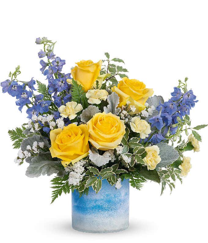 A floral arrangement featuring yellow roses, light yellow mini carnations, blue delphinium, white statice, and floral greens in a frosted blue skies cylinder vase.