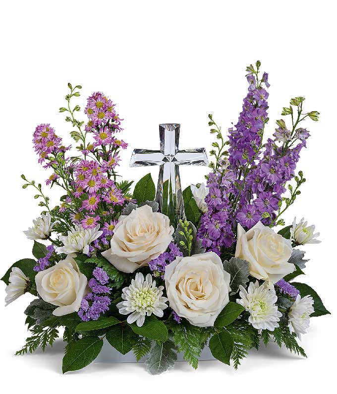 A graceful floral arrangement showcasing crme roses, lavender larkspur, lavender asters, lavender statice, white mums, and floral greens, elegantly accented with a crystal cross. This bouquet blends soft, calming colors for a serene display.