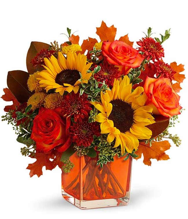 A vibrant floral arrangement featuring orange roses, yellow sunflowers, gold cushion poms, and rust cushion mums, accented with seeded eucalyptus, oregonia, and magnolia leaves, all beautifully presented in a keepsake orange cube vase.