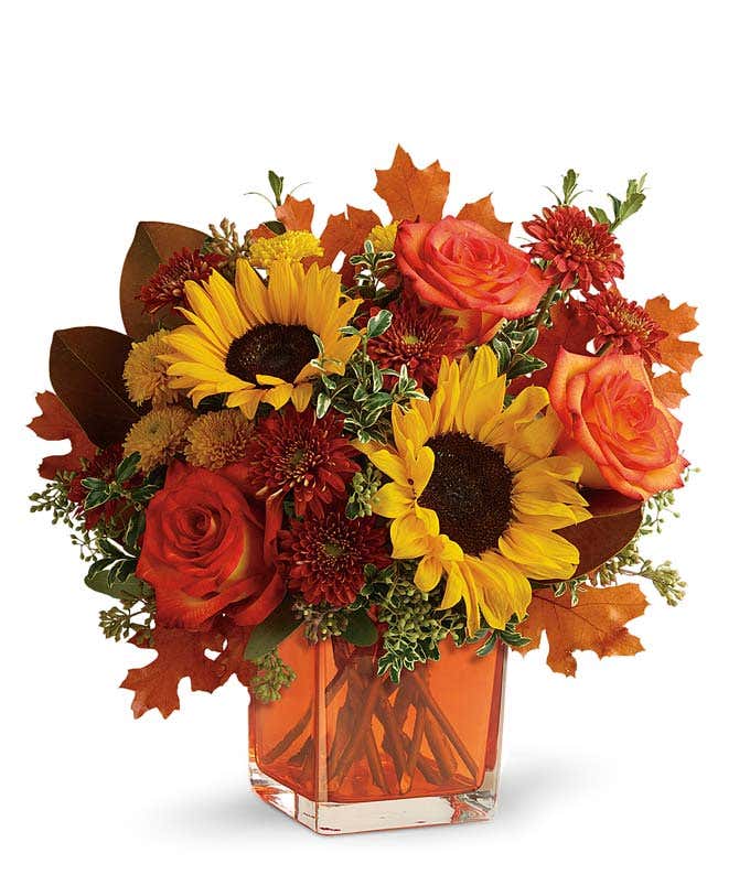 A vibrant floral arrangement featuring orange roses, yellow sunflowers, gold cushion poms, and rust cushion mums, accented with seeded eucalyptus, oregonia, and magnolia leaves, all beautifully presented in a keepsake orange cube vase.