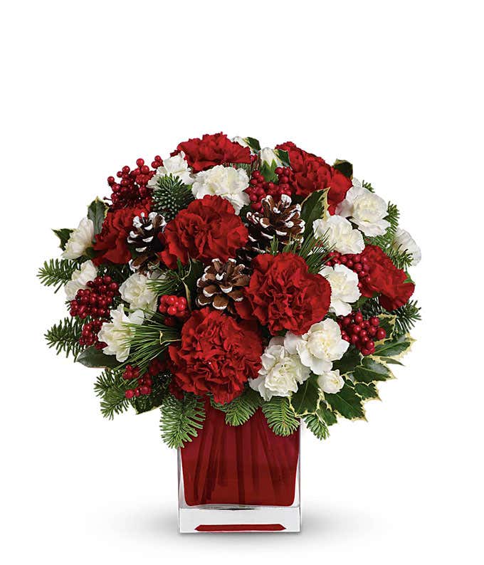 A floral arrangement featuring vibrant red carnations and delicate white mini carnations, complemented by lush noble fir and white pine, all elegantly displayed in a red glass cube vase.