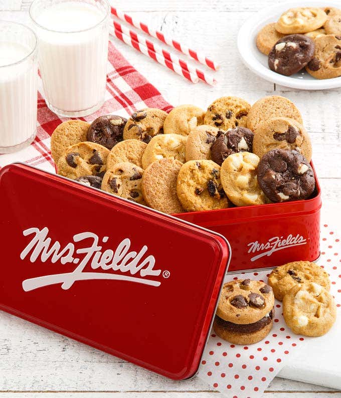  Red Mrs. Fields tin filled with a variety of mini cookies, including chocolate chip, white chocolate macadamia, and double chocolate, displayed with glasses of milk and striped straws on a white wooden surface with a red checkered napkin.