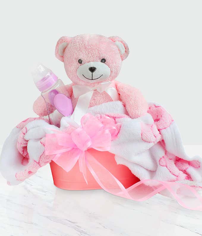 A pink baby gift basket with a teddy bear, baby bottle, and soft pink and white blanket, adorned with a pink ribbon.