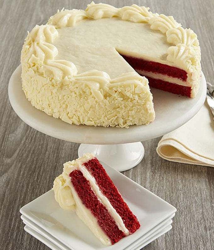 A red velvet cake with white cream cheese frosting, garnished with white chocolate shavings, displayed on a white cake stand. A slice of the cake is served on a small white plate, showcasing the rich red interior and creamy layers.